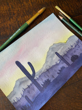 Load image into Gallery viewer, Painting of a desert landscape with cacti on a wooden surface with art supplies.