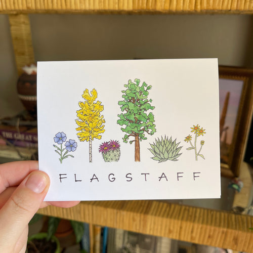Hand holding a notecard with watercolor illustrations of plants, flowers, and cacti, and 'Flagstaff' at the bottom.