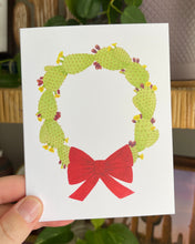 Load image into Gallery viewer, Hand holding a white notecard with a watercolor illustration of a green wreath made of prickly pears and red bow.