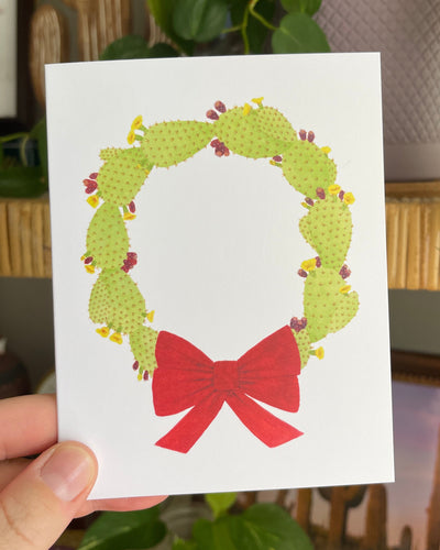 Hand holding a white notecard with a watercolor illustration of a green wreath made of prickly pears and red bow.