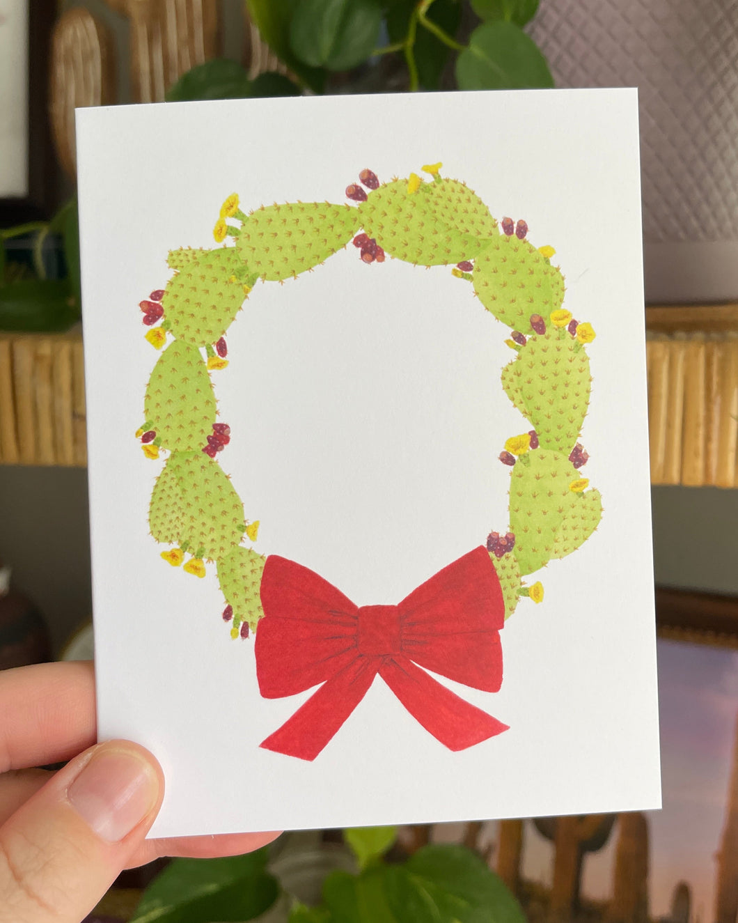 Hand holding a white notecard with a watercolor illustration of a green wreath made of prickly pears and red bow.