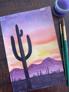 A watercolor illustration of a desert scene at sunset, in the foreground is a large saguaro cactus with 3 arms, the sky is a brilliant purple, orange, pink, and yellow at sunset, with purple mountains in the background. Sitting on the wooden table next to this watercolor painting is a small cup of water and two paintbrushes. 