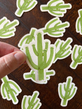 Load image into Gallery viewer, Hand holding a green saguaro sticker with more saguaro stickers on a wooden surface