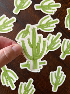Hand holding a green saguaro sticker with more saguaro stickers on a wooden surface