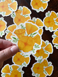 Hand holding a sticker with a watercolor illustration of orange poppy flowers with more stickers in the background on a dark wooden table