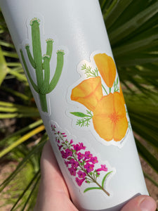 A hand holding a white waterbottle with 3 stickers on it, one of a green saguaro, one of a pink penstemon, and one of orange poppy flowers, with green palm tree leaves in the background.