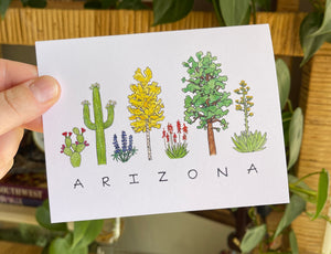 Arizona Flora greeting card, the word "ARIZONA" written in all caps handwriting with an assortment of plants painted in watercolor above. Including prickly pear, saguaro, lupine, aspen, elf aloe, ponderossa pine, and agave.
