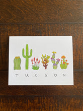 Load image into Gallery viewer, Tucson Flora greeting card, the word &quot;TUCSON&quot; written in all caps handwriting with an assortment of plants painted in watercolor above. Including barrel cactus, saguaro, elf aloe, teddy bear cholla, prickly pear, and torch cactus.