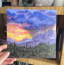 Load image into Gallery viewer, Hand holding a print of a watercolor painting showing a vibrant sunset sky with rain from a monsoon pouring over the desert