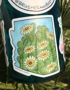 Vinyl sticker in the shape of Arizona with a watercolor painting of the top of a saguaro cactus with white blooming flowers. The sticker is on a water bottle with other stickers by Brushes and Boots.